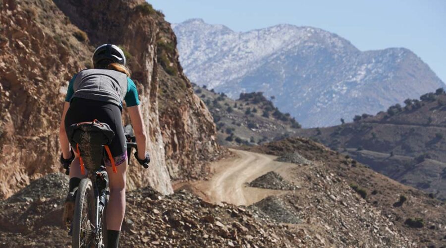 A mountain biker in professional gear ascends a gravel path in the Atlas Mountains, with snow-capped peaks in the backdrop under a clear sky.