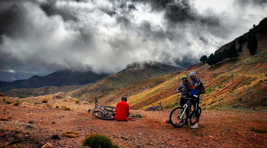 Two mountain bikers and a bike laying on its side rest during a cloudy day on the rugged slopes of the Atlas Mountains, showcasing the dynamic terrain and moody skies.