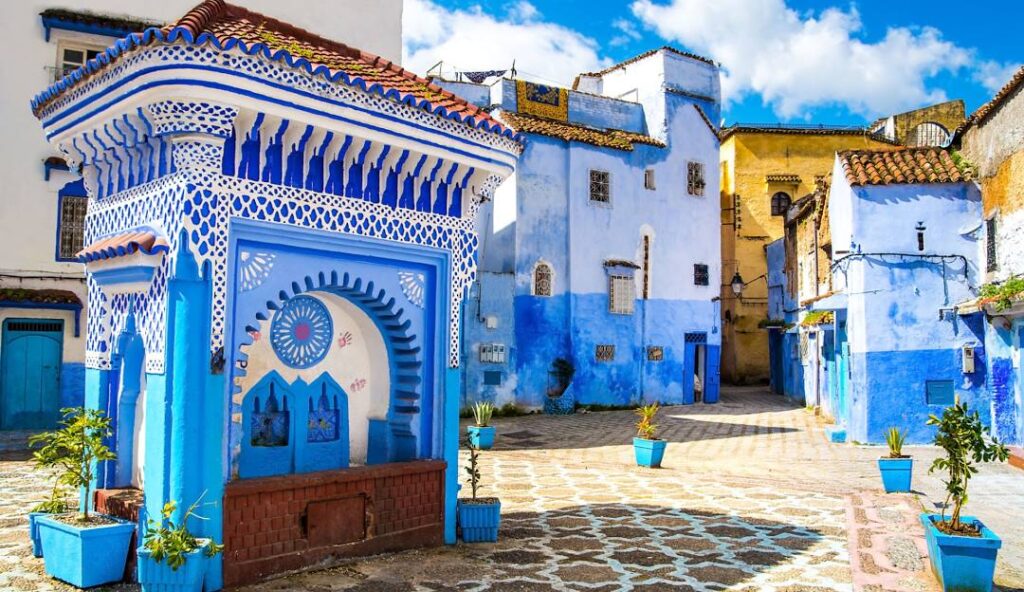 A cobblestone square in Chefchaouen with a striking blue and white traditional Moroccan building adorned with intricate patterns, surrounded by houses with blue walls and terracotta roofs under a sunny sky.