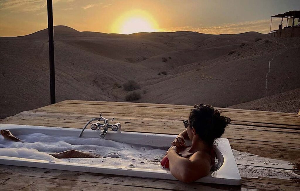 Person relaxing in an outdoor bathtub with a view of the sun setting over Agafay Desert dunes.
