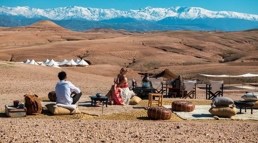 A couple enjoys a scenic view of the Agafay Desert with luxurious glamping tents and the snow-capped Atlas Mountains in the background, preparing for a sunset camel ride and Moroccan dinner.