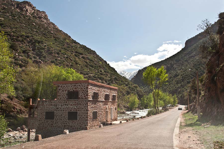 Rural road in Ourika Valley with traditional stone building and Atlas Mountains in the distance.