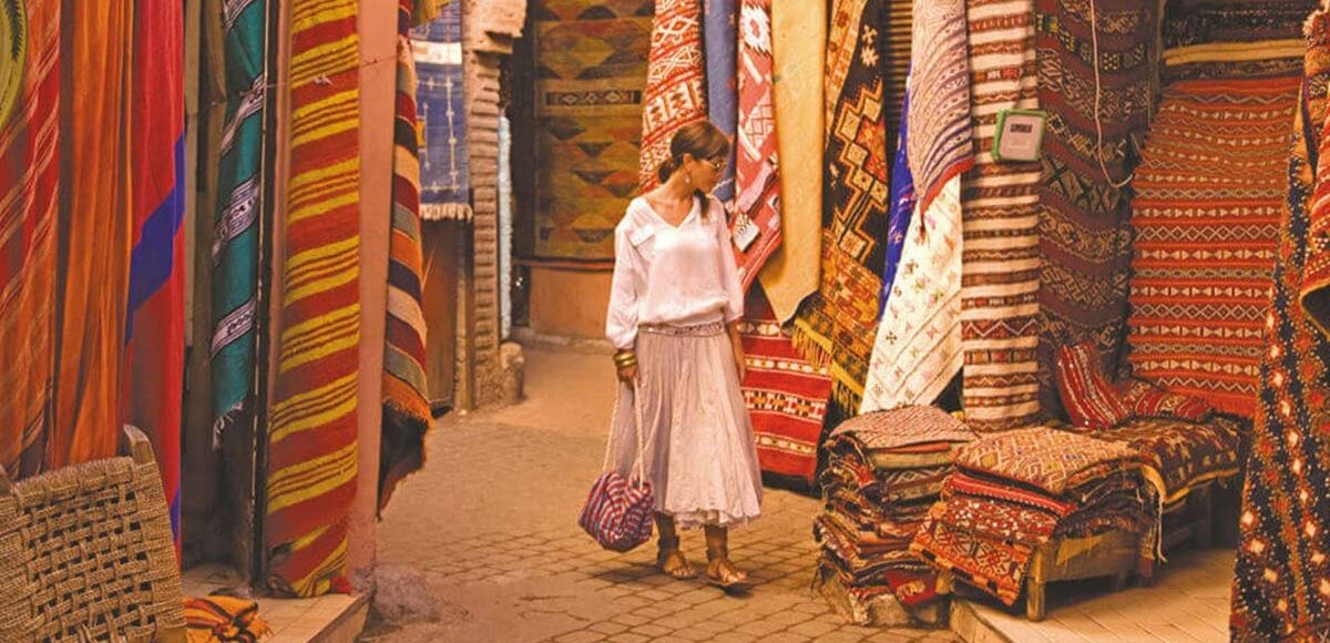 Woman in white blouse and skirt walking amidst vibrant, colorful traditional rugs and textiles displayed on walls and piles in a Marrakech souk, showcasing the rich culture and craftsmanship of Moroccan markets.