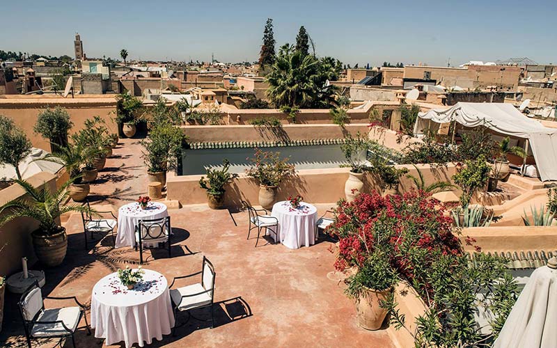 Spacious rooftop terrace of a Moroccan riad, featuring elegant dining setups and lush plants, with a view of Marrakech’s skyline.