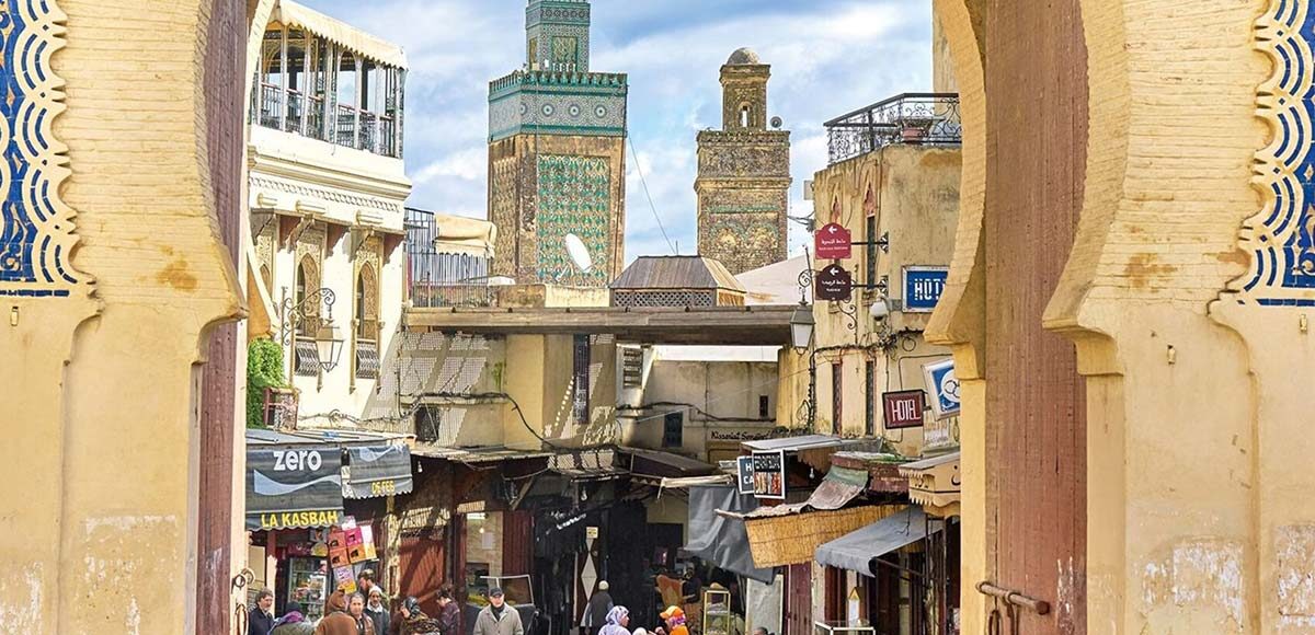 Bab Bou Jeloud (Blue Gate) in Fes, Morocco, framing the bustling street of the medina with shops, cafes, and the green-tiled minaret in the background.