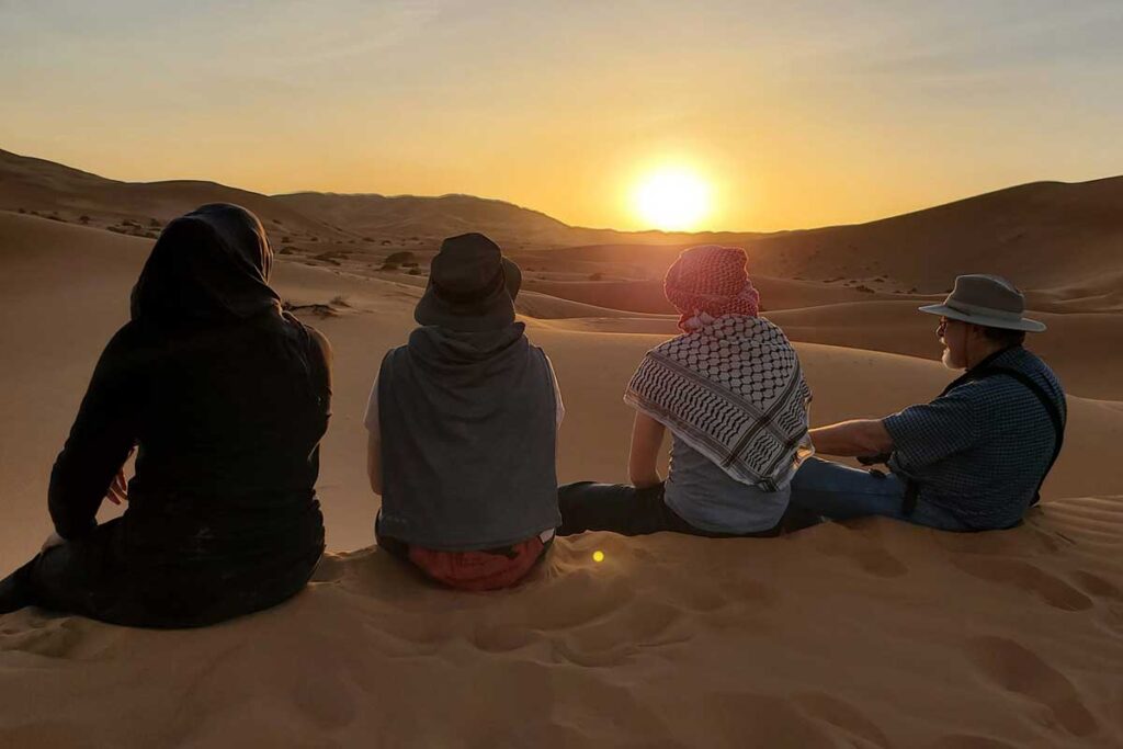 Travelers sitting on Erg Chebbi dunes watching sunset in Merzouga Sahara Desert.