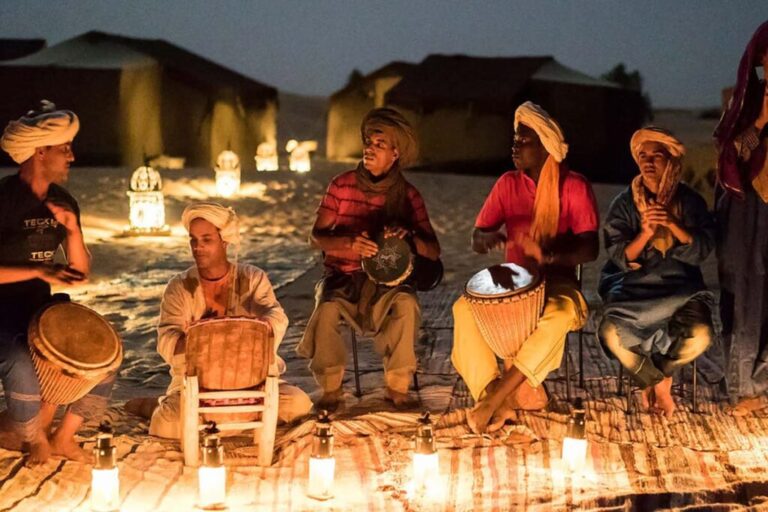 Berber musicians playing drums and percussion by candlelight at a Merzouga desert camp at night, with traditional tents visible in the background