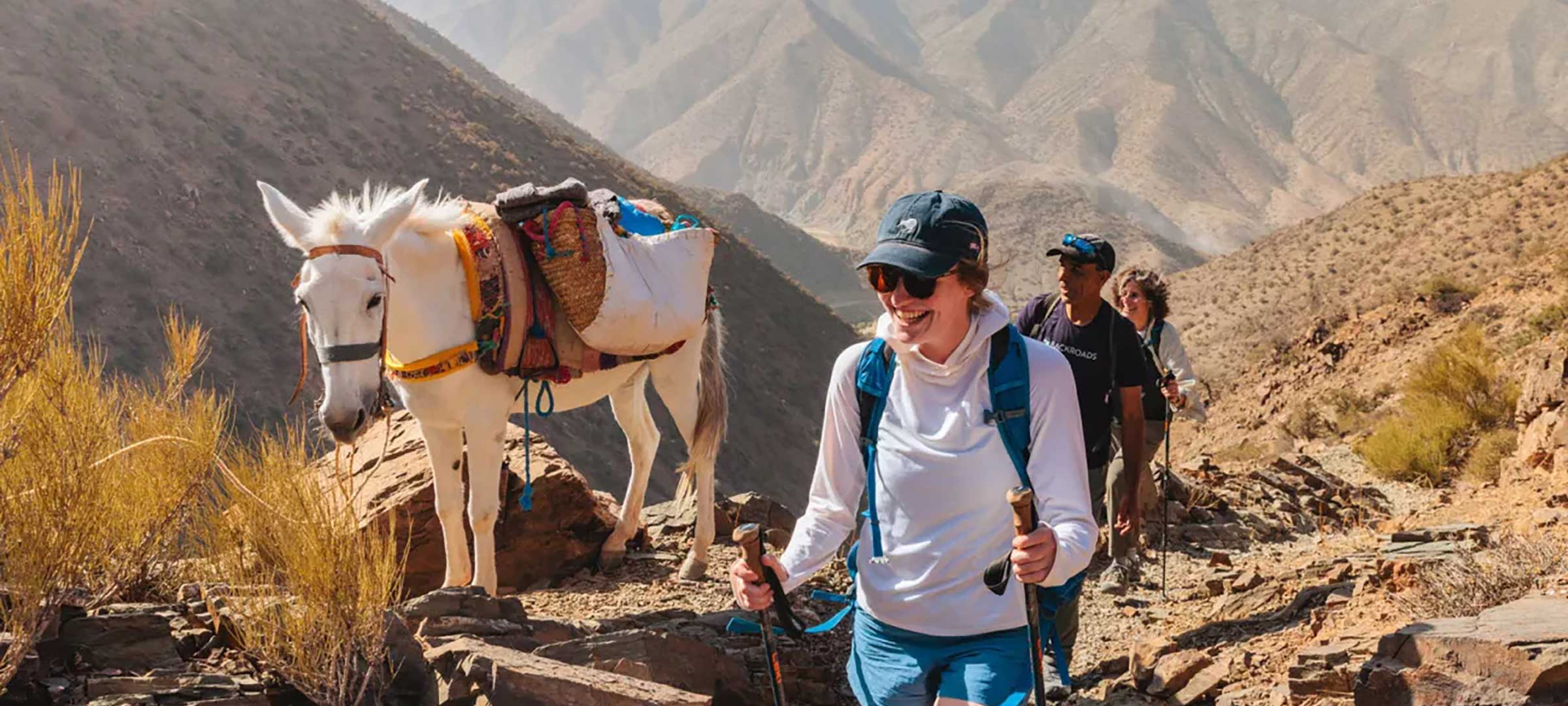 Smiling hiker with trekking poles walking beside a white pack mule on a 3-Day Atlas Mountains Trek, guide and trekker behind in the High Atlas