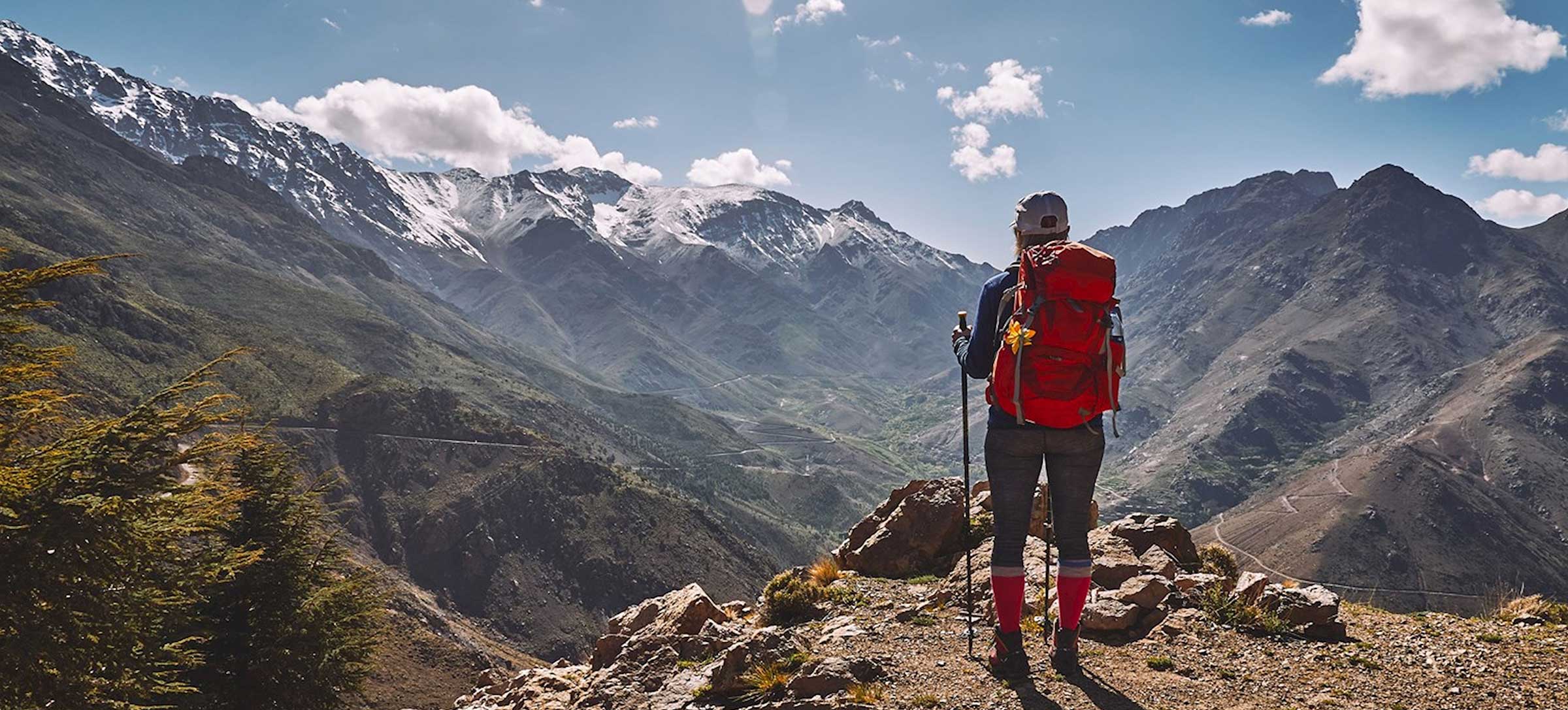 Woman hiker with red backpack viewing snow-capped High Atlas peaks on the Imlil to Ourika 3-day trek