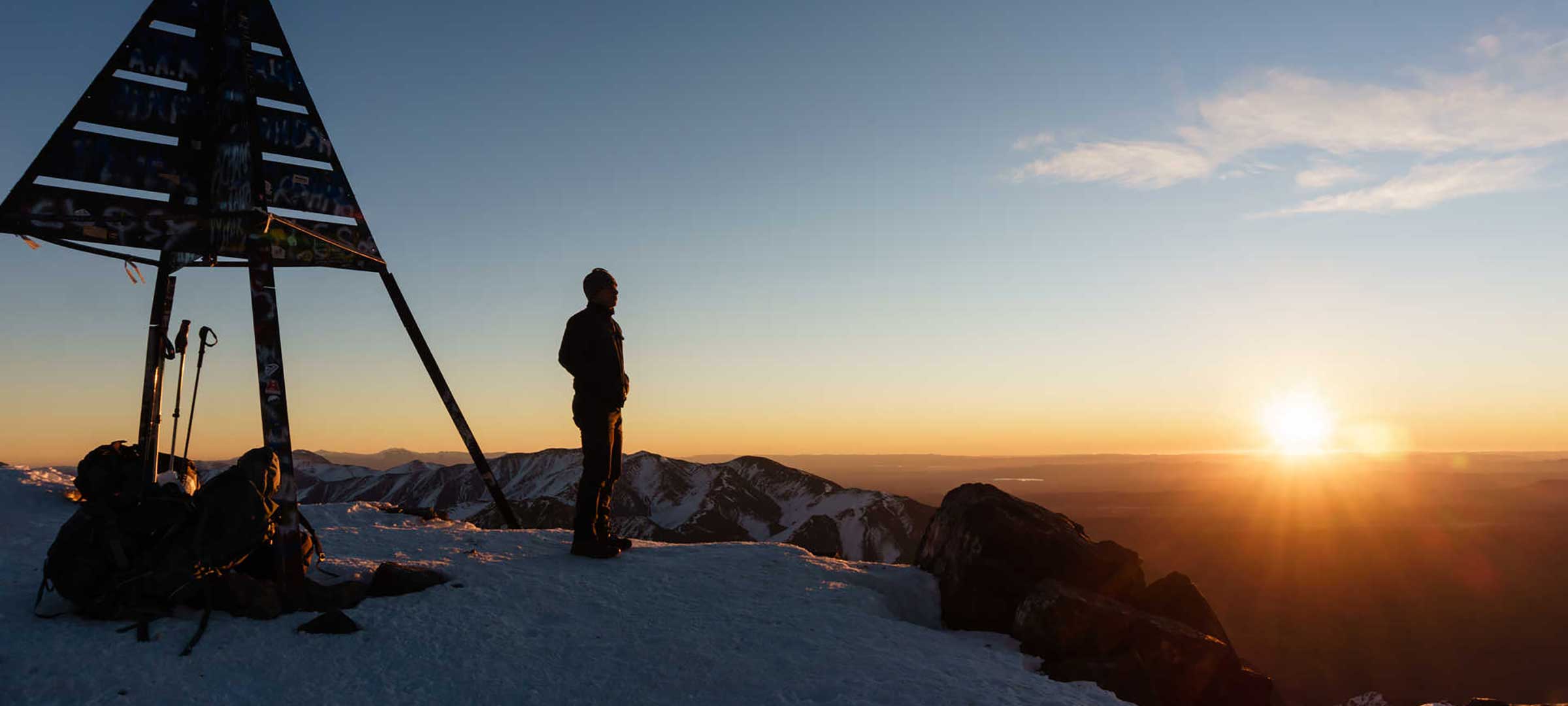 Trekkers at the Mount Toubkal summit marker at 4,167m watching sunrise over the Atlas Mountains in winter