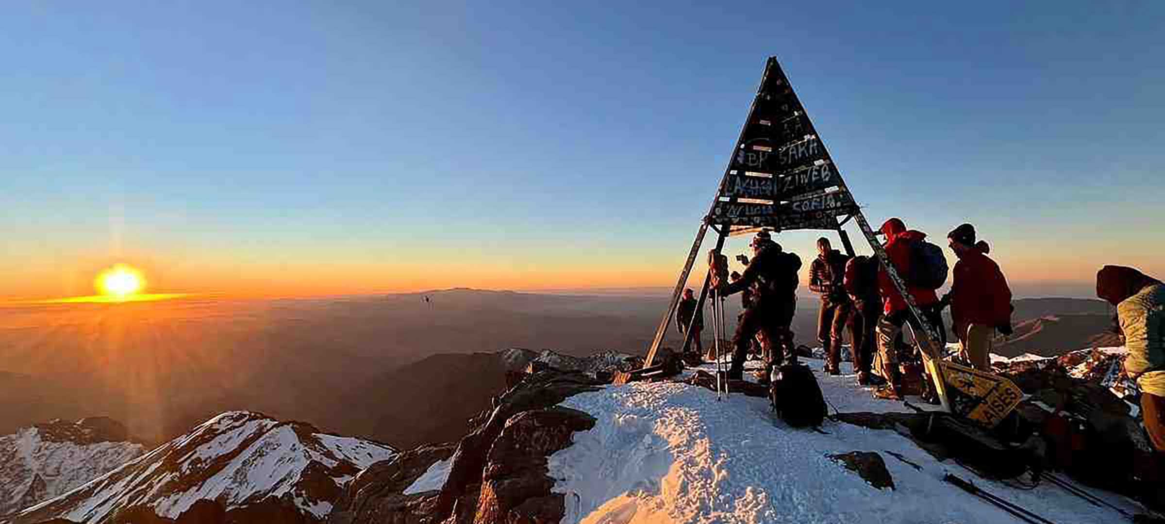 Trekkers at the Mount Toubkal summit marker at 4,167m watching sunrise over the Atlas Mountains in winter