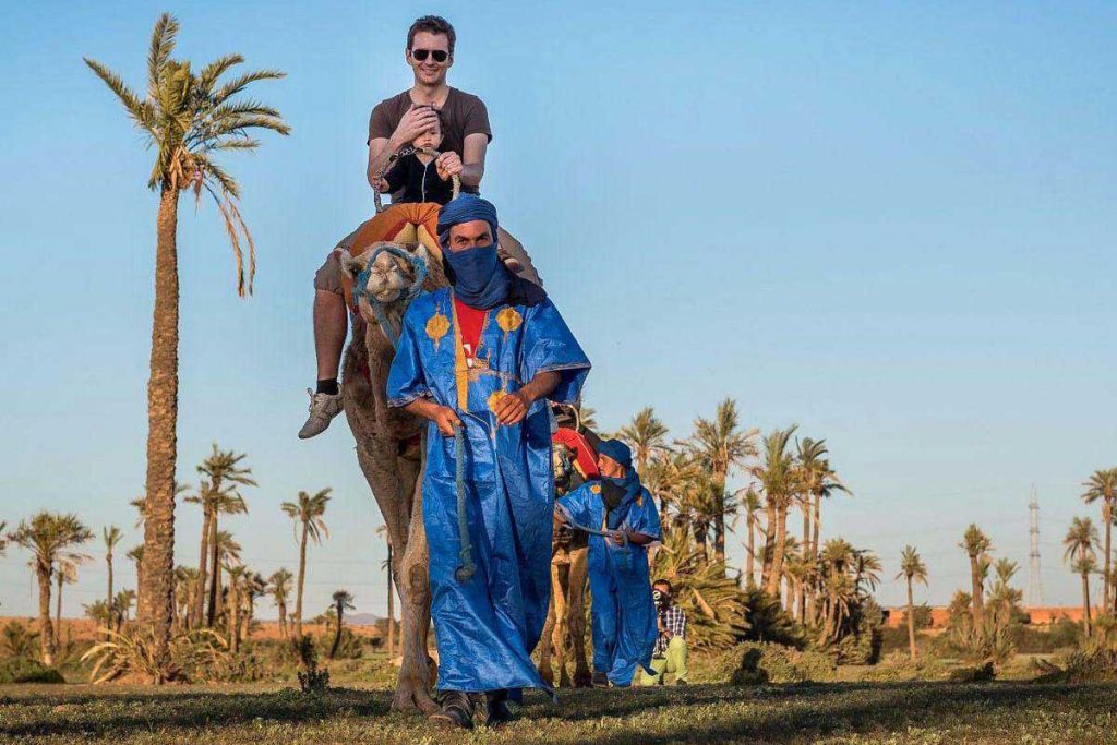 Père et jeune enfant à dos de chameau dans la palmeraie de Marrakech, guidés à pied par deux Berbères en tenue saharienne bleue
