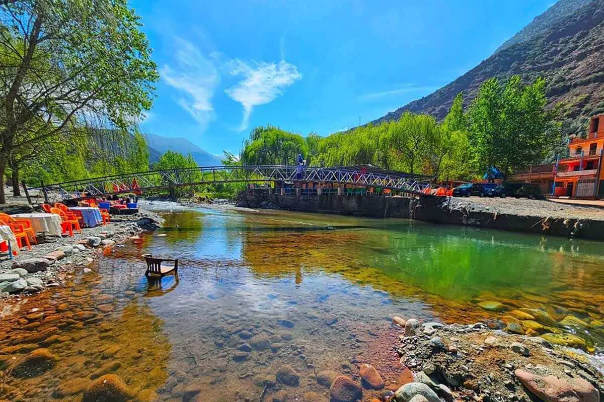Wooden footbridge over the clear Ourika River with riverside café chairs on the bank, day trip from Marrakech