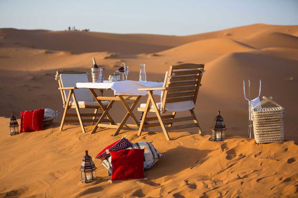 Romantic private dinner table set in the Erg Chebbi dunes at sunset, with champagne, lanterns and Berber cushions, on a 3-day Marrakech to Merzouga private desert tour