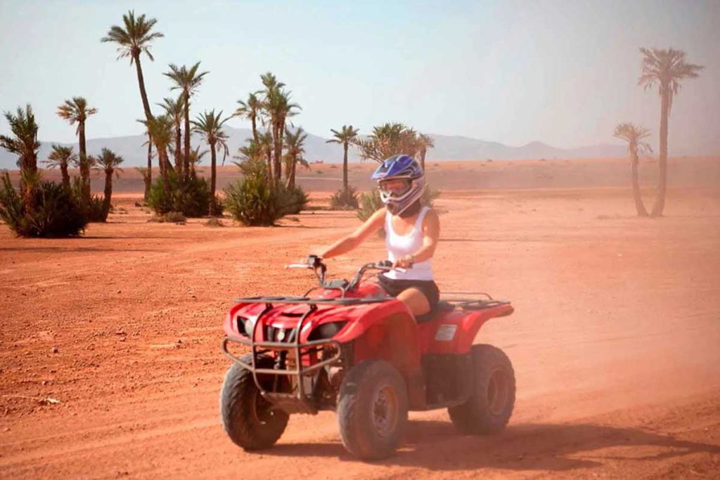 Woman riding a red quad bike across the red-earth tracks of the Marrakech Palmeraie, palm trees and Atlas foothills behind