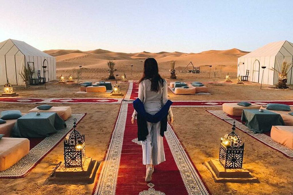 Woman walking a red carpet path into a luxury desert camp lit by Moroccan lanterns at dusk in the Erg Chebbi dunes near Merzouga