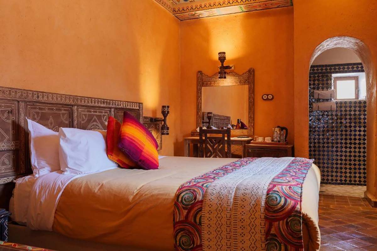 Guest room at Kasbah Hotel Tomboctou with a carved wooden headboard, ochre walls, colourful Berber throw and zellige en-suite