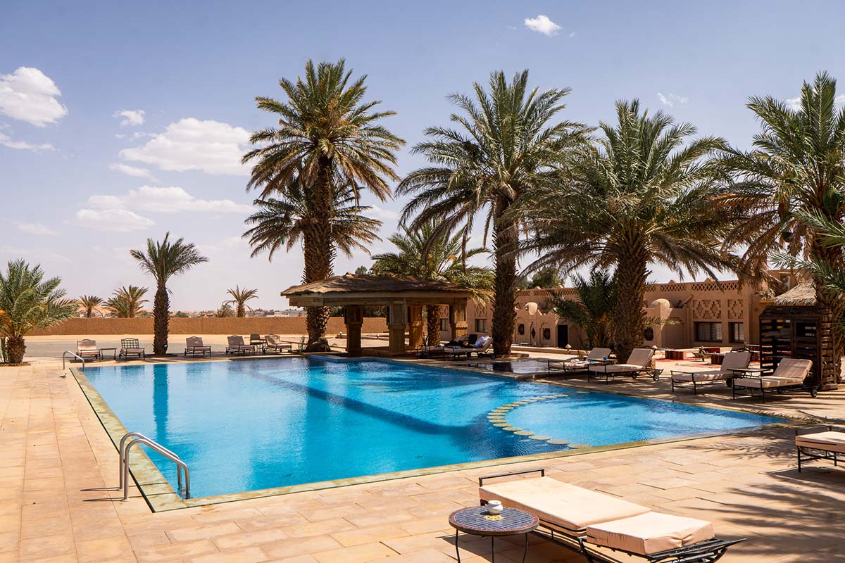 Large swimming pool at Kasbah Hotel Tomboctou with a thatched pavilion, tall date palms, sun loungers and desert views