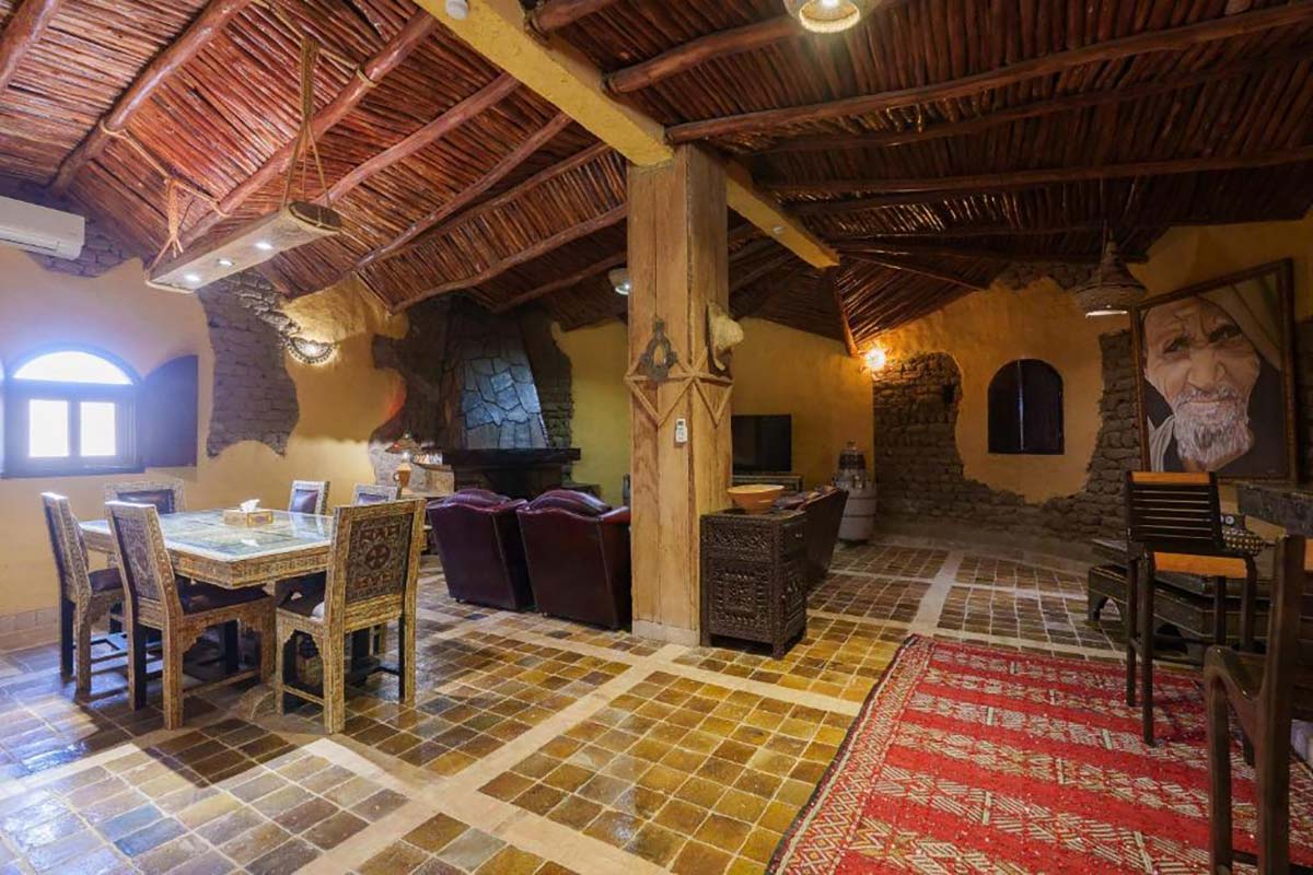Lounge and dining room at Kasbah Hotel Tomboctou with a reed-and-beam ceiling, stone fireplace, tiled table and Berber rug