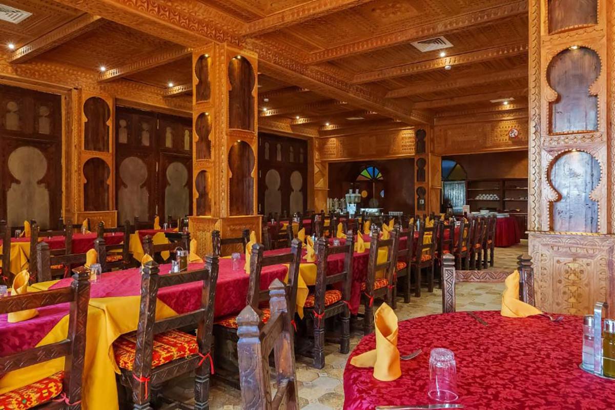 Restaurant dining hall at Kasbah Hotel Tomboctou with carved cedar pillars, arched panelling and red-and-yellow set tables